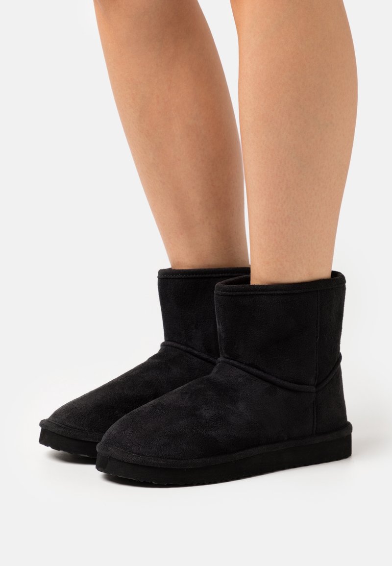 Bottes mi-mollet en daim noir portées sur des jambes nues, avec des bouts arrondis et des semelles en caoutchouc plates sur fond blanc.