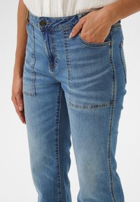 Modre denim hlače z visokim pasom, sprednjimi žepi, diskretnim bledenjem in kontrastnim šivanjem ob šivih. Gladek tekstura.