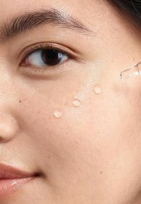 Des gouttes de sérum de soin de la peau transparent sont présentées sur une peau lisse et claire. Un compte-gouttes dispense une autre goutte près de l'os de la joue.