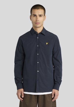 Jeune homme aux cheveux courts porte une chemise bleu foncé boutonnée avec un logo d'oiseau jaune sur la poitrine et un pantalon marron sur fond gris.