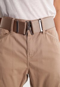 Khaki pasek z teksturowanej tkaniny, z metalową klamrą i zapięciem w kolorze srebrnym. Nosi się go na beżowych spodniach o gładkim wykończeniu.