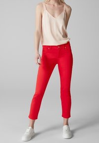 Jean fitted rouge avec une texture lisse et un design à cinq poches, associé à un débardeur en satin rose clair et des baskets blanches.