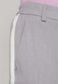 Grå byxor med en slät textur, med en vit sidremsa, elastisk midjeband och sidofickor. Diskret varumärkesskylt synlig.