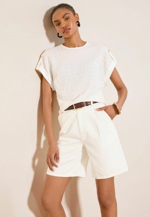 Junge Frau trägt weißes strukturiertes kurzärmeliges Oberteil, hoch taillierte weiße Shorts mit braunem Gürtel, posiert mit einer Hand in der Tasche vor einer beigen Wand.
