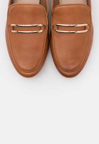 Mocassins en cuir brun avec une texture lisse, un bout rond et une barre métallique sur le devant. La semelle est légèrement surélevée.