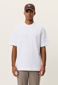 Λευκό βαμβακερό t-shirt με στρογγυλή λαιμόκοψη και κοντά μανίκια, συνδυασμένο με καφέ καπέλο με λογότυπο, που φοριέται από ένα άτομο σε όρθια στάση.