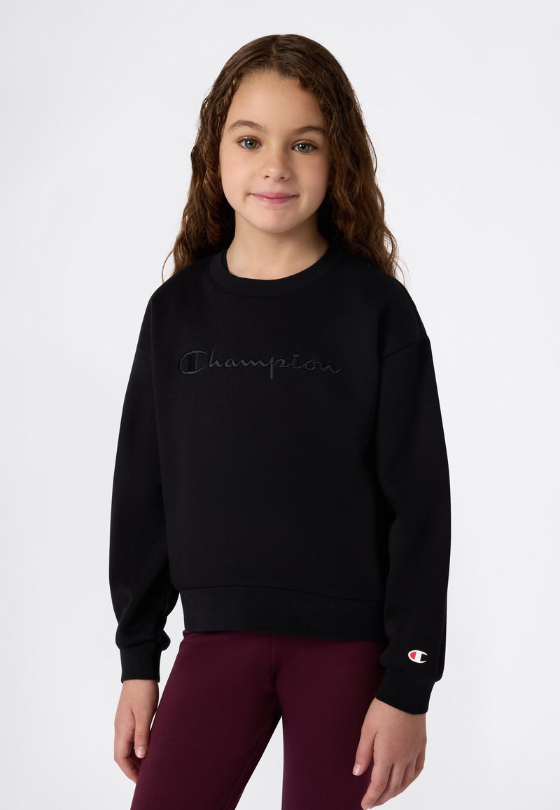 Ragazzina con lunghi capelli ricci che indossa un sweatshirt nero della Champion e pantaloni bordeaux, in piedi contro uno sfondo bianco semplice.