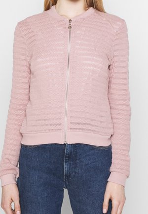 Kobieta w jasnoróżowym, teksturowanym swetrze na zamek z ściągaczami przy rękawach i niebieskich dżinsach, przedstawiona od szyi do bioder na jednolitym tle.