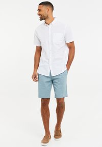 Bílá košile s krátkým rukávem a náprsní kapsou v kombinaci se světle modrými šortkami a hnědými nazouvacími botami. Jednoduchý, neformální design.