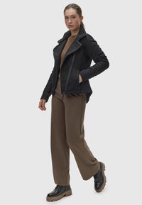 Giacca nera in tessuto testurizzato, con colletto in pelliccia sintetica, chiusura con zip e tasche laterali. Un maglione marrone, pantaloni beige e stivali neri chunky completano il look.
