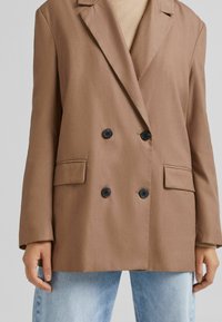 Hnědý dvouřadý blazer z hladké látky s černými knoflíky, dvěma předními kapsami a klopami s výřezem, doplněný světle modrými džíny.