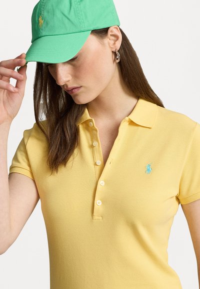 Rumena polo majica s kratkimi rokavi in gumbi, z našitim turkiznim logotipom. Zelen klobuk nošen pod kotom.