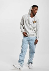 Grå zip-up hettegenser med en frontgrafikk, kombinert med lyseblå jeans og hvite sneakers. Hettegenseren har snor i hetten og ribbestrikkede ermer.