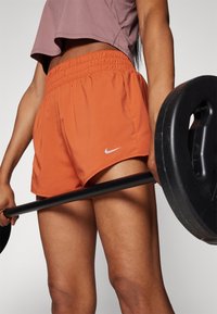 Shorts deportivos naranjas con una cinturilla elástica y un pequeño logo plateado de Nike, combinados con una camiseta corta morada, sosteniendo una barra de pesas.
