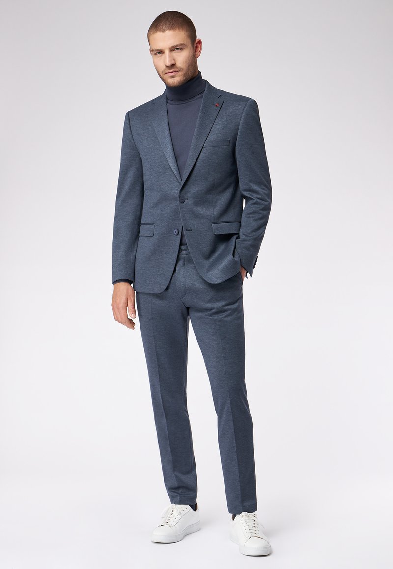 Completo da uomo in blu scuro, realizzato in tessuto strutturato, composto da un blazer monopetto e pantaloni slim-fit. Indossato con un dolcevita scuro e sneakers bianche.