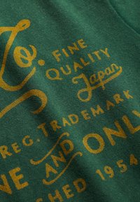 Tela verde con texto impreso en dorado que dice "Fine Quality Japan" y "Established 1954", mostrando una textura suave y letras curvas.