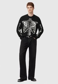 Must kampsun, millel on valge rinnakorvi muster, koos lahtiste mustade teksade ja mustade kingadega. Silmapaistev tekstuur, vabalt istuv lõige ja graafiline detail.