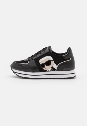 Zapatilla negra con detalles brillantes, suela blanca y cara de dibujo animado con gafas de sol en el lateral, mostrada de perfil izquierdo.