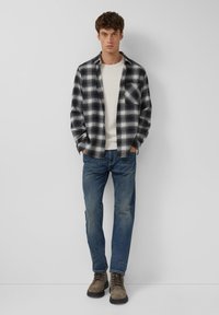 Jonge man staat tegen een witte muur, gekleed in een zwart-wit geruite overhemd, een crème sweater, blauwe jeans en bruine veterschoenen.