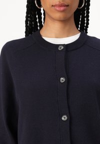 Námořnický modrý cardigan s obláčkovým výstřihem, s knoflíkovým zapínáním, žebrovanými okraji a hladkou texturou. Dva knoflíky jsou viditelné na vrchu.