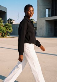 Jonge zwarte vrouw die buiten loopt in een witte broek en een zwart jasje, met moderne betonnen gebouwen en palmbomen op de achtergrond.