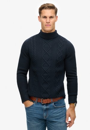 Suéter de cuello alto acanalado en color azul marino con diseño de punto trenzado, con mangas largas y una silueta ajustada, combinado con jeans de color azul claro y un cinturón marrón.