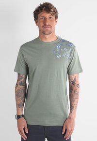 T-shirt en coton vert olive avec un graphique floral bleu sur l'épaule gauche. Manches courtes, col rond, texture douce.
