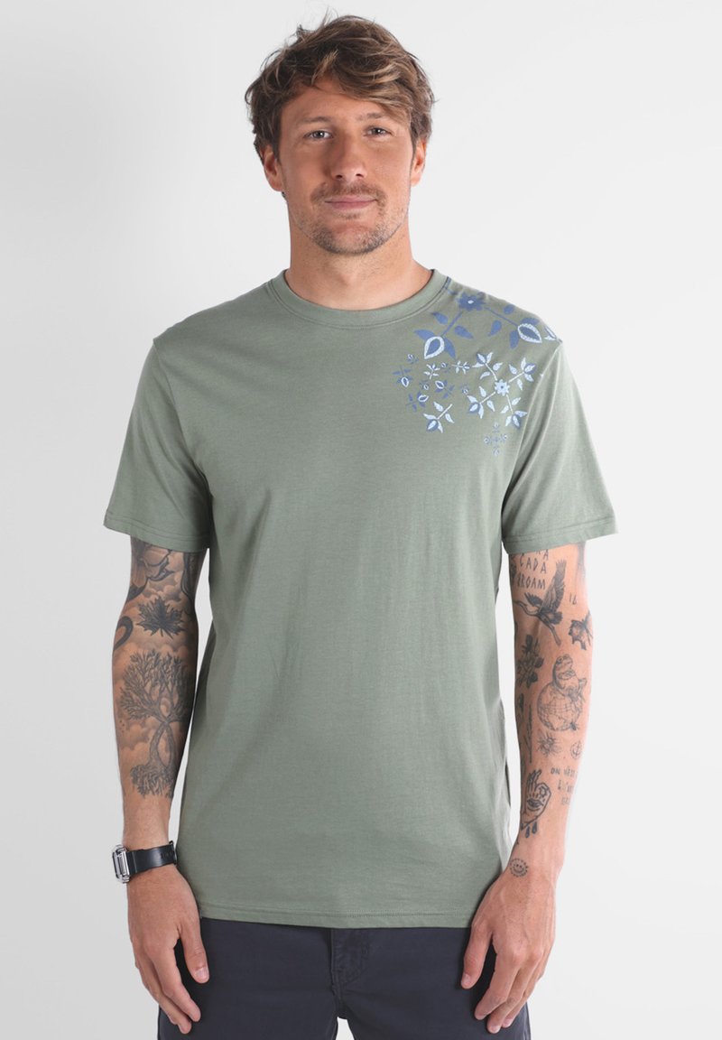 T-shirt en coton vert olive avec un graphique floral bleu sur l'épaule gauche. Manches courtes, col rond, texture douce.