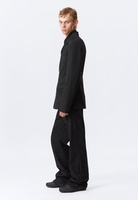 Svart pinstripet dress med en skreddersydd blazer og løstsittende bukser. Materialet ser glatt ut med en lett glans; teksturerte gummisålte sko.