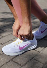 Zapatilla deportiva blanca con acentos morados y amarillos, parte superior texturizada, diseño de cordones y amortiguación visible, usada en un camino de adoquines.