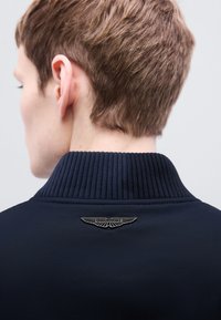 Μπλε μπουφάν με ριμπ γιακά και ασημένιο λογότυπο Aston Martin στην πλάτη, με λεία υφή και κομψό, αεροδυναμικό σχέδιο.