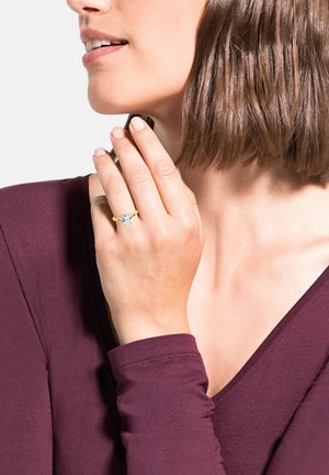 Anello d'oro con un grande diamante tondo, caratterizzato da una fascia gialla liscia. La modella indossa una maglietta di un profondo bordeaux, mettendo in mostra l'anello sulla sua mano.