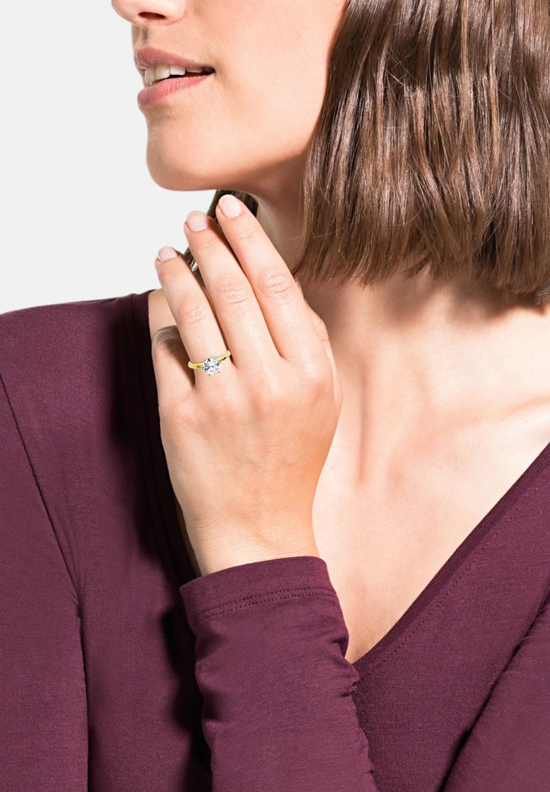 Anello d'oro con un grande diamante tondo, caratterizzato da una fascia gialla liscia. La modella indossa una maglietta di un profondo bordeaux, mettendo in mostra l'anello sulla sua mano.