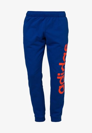 Pantalon de jogging bleu en tissu doux, avec un logo "adidas" orange imprimé verticalement sur le côté gauche et une taille élastique.
