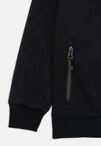 Section de veste noire avec une texture lisse, présentant des poignets côtelés et une poche zippée avec des accents métalliques sur la manche gauche.