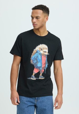 Junger Mann trägt ein schwarzes T-Shirt mit einer Grafik eines Igels in einer blauen Jacke, roten Hosen und Turnschuhen, der vor einem schlichten Hintergrund steht.