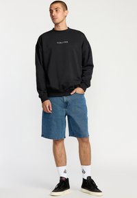 Black sweatshirt with "VOLCOM" logo, blue denim shorts, white socks with logo, and black sneakers. Casual outfit with relaxed fit.