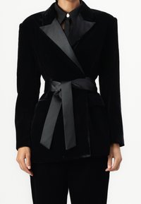 Czarna aksamitna marynarka z satynowym paskiem. Posiada kołnierz szalowy i dwie przednie kieszenie. Gładka textura z eleganckim, dopasowanym krojem.