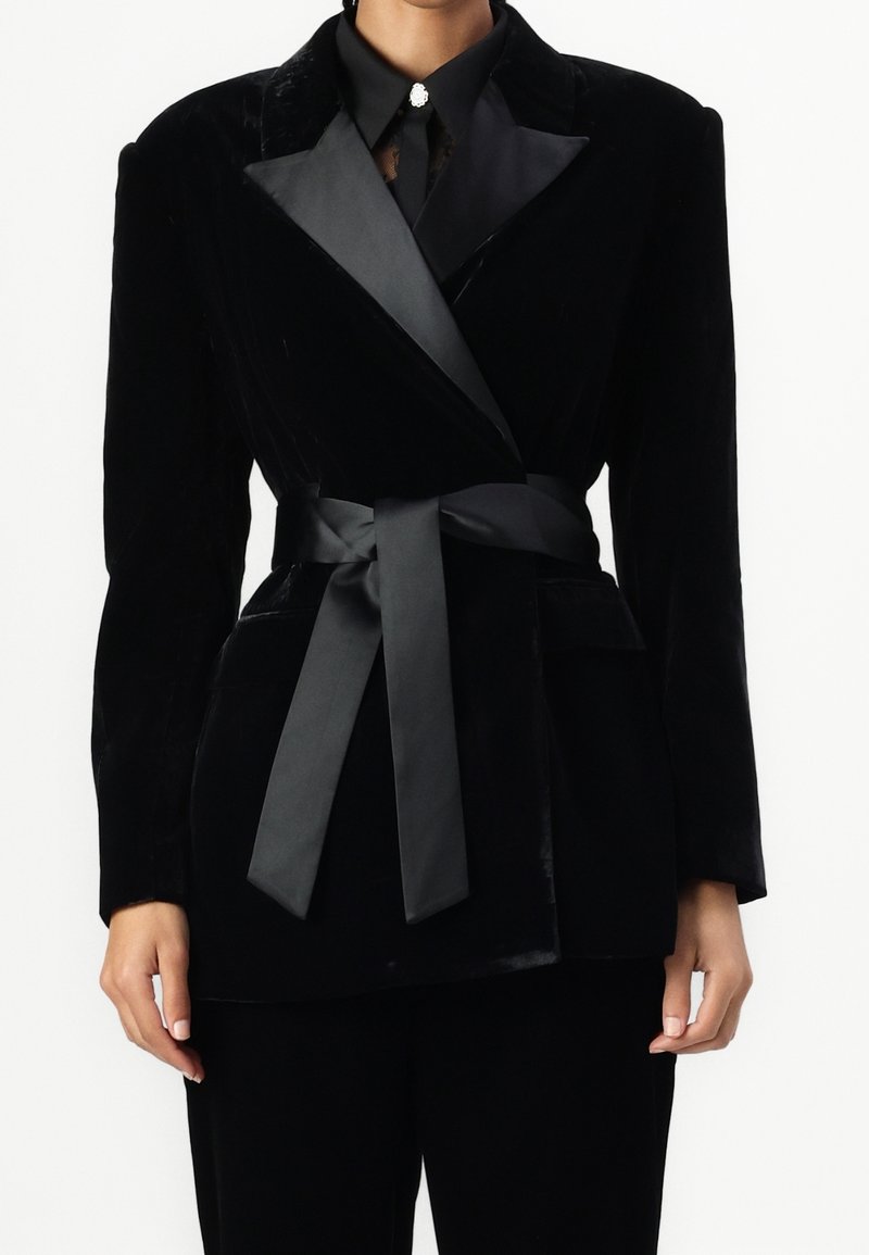 Czarna aksamitna marynarka z satynowym paskiem. Posiada kołnierz szalowy i dwie przednie kieszenie. Gładka textura z eleganckim, dopasowanym krojem.