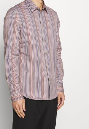 Man wearing pastel-colored vertical striped button-up shirt with long sleeves and black pants against a plain light background.
