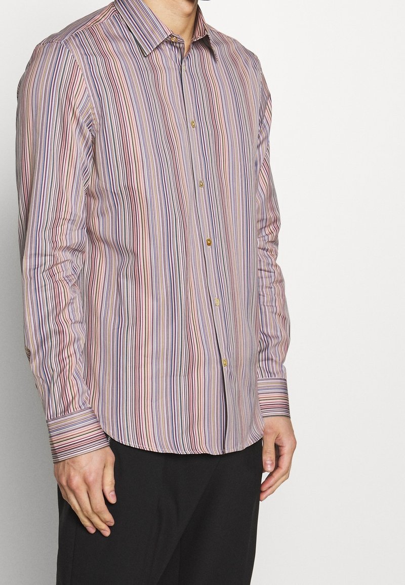 Homme portant une chemise à boutons à rayures verticales de couleurs pastel avec manches longues et un pantalon noir, sur un fond clair uni.