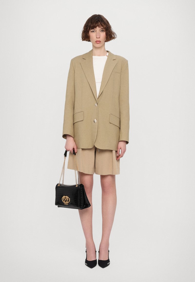 Blazer beige oversize con due tasche frontali, indossato sopra un top chiaro e pantaloni corti beige. Borsa nera con logo dorato, abbinata a tacchi neri.