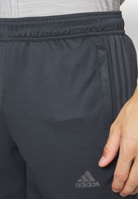 Czarne spodnie sportowe z elastycznym pasem, teksturowana tkanina, boczne kieszenie i trzy pionowe paski. Zawierają logo Adidas w odcieniu szarości.