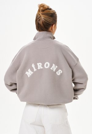 Frau mit Dutt trägt eine lockere graue Jacke mit hohem Kragen und weiße Hose. Die Jacke hat "MÍRONS" in weißen Buchstaben auf dem Rücken.