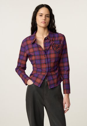 Vrouw draagt een rood en paars geruit overhemd met knopen en een hartvormige patch en zwarte broek, staand met één hand in de zak.