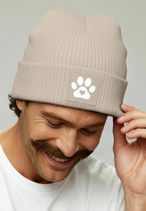 Man with mustache smiling, adjusting beige ribbed beanie featuring a white paw print with a heart in the center patch.