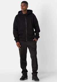Sudadera negra con cremallera fabricada en un tejido suave, con un bolsillo frontal, puños acanalados y capucha. Combinada con pantalones negros y zapatillas.