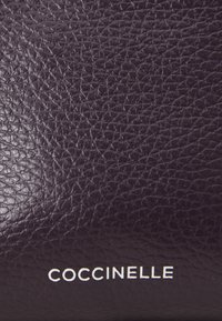 Lila bőrtáska texturált felülettel és finom erezettel; a "Coccinelle" márkanevével fehér betűkkel.