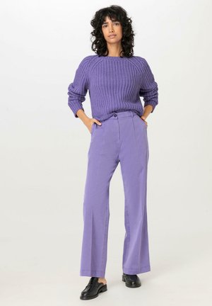 Maglione viola lavorato a maglia con texture a coste, abbinato a pantaloni viola a gamba dritta e a vita alta. Scarpe nere senza lacci completano l'outfit.