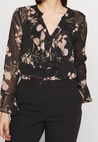 Svart blommig blus med långa, genomskinliga ärmar, volangkrage och rosa blommönster. Kombinerad med högmidjade svarta byxor.
